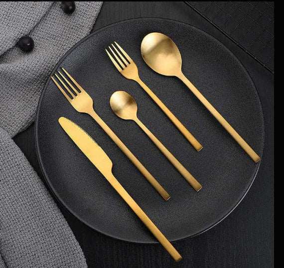Matte stainless steel cutlery set with gold finish on black grid mat, includes knife, forks, and spoons.